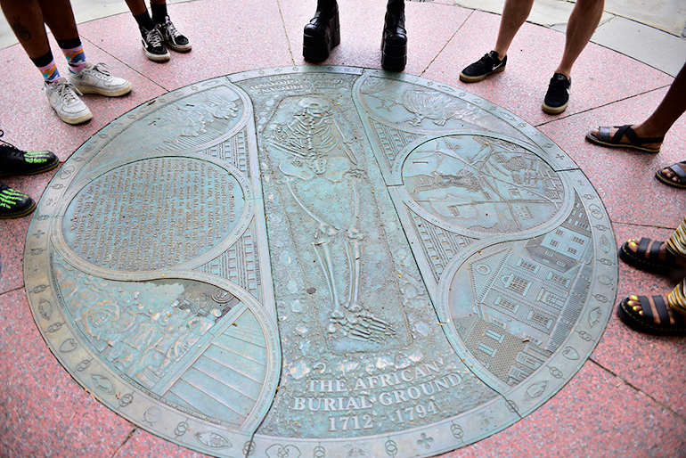 Participants circle the bronze medallion for the African Burial Ground. In the center of the Medallion is a skeleton and broken shackles.