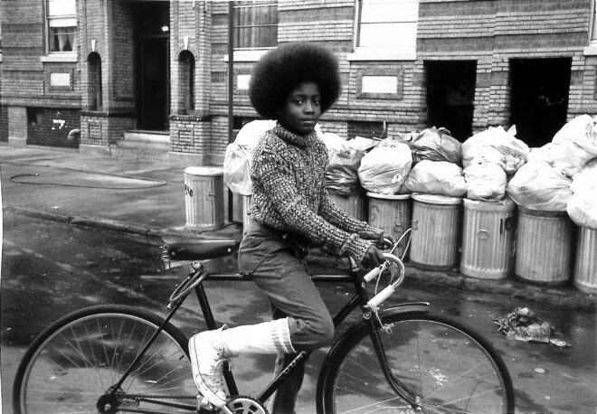 Arlene Gottfried (1950–2017) Boy with Afro on Bicycle in front of Trash Cans, 1975 Gelatin silver print The New York Historical, Gift of Sally Klingenstein Martell © Estate of Arlene Gottfried.